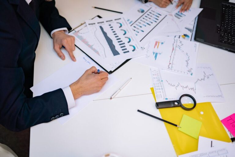 Two professionals review charts and graphs spread across a white desk, with a laptop nearby and sticky notes visible on the side.