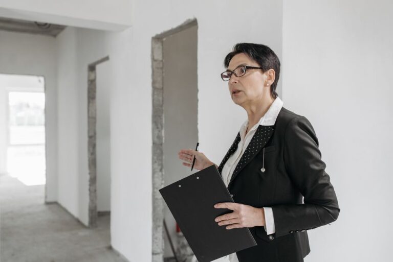 Professional woman in a suit with a clipboard speaks during a site visit in an unfinished white-walled interior.