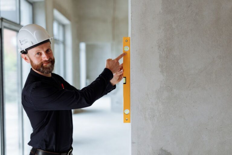 Construction worker in a white hard hat uses a yellow spirit level to check the wall's alignment in a concrete interior.