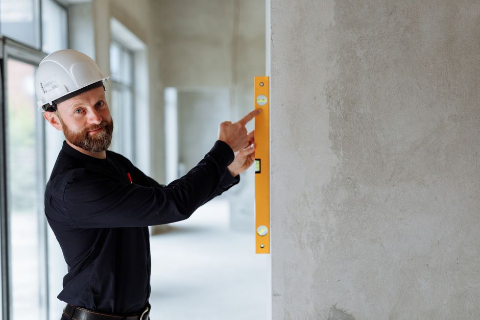 Construction worker in a white hard hat uses a yellow spirit level to check the wall's alignment in a concrete interior.