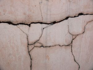 Close-up of a beige concrete wall with deep cracks and jagged joints throughout the surface, showing weathering.