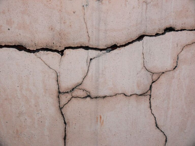 Close-up of a beige concrete wall with deep cracks and jagged joints throughout the surface, showing weathering.