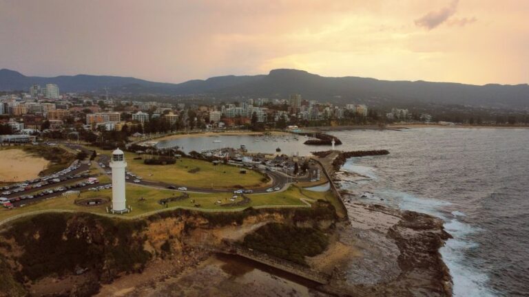 A coastal town at sunset with a white lighthouse on a grassy cliff overlooking a harbor and rocky shoreline.