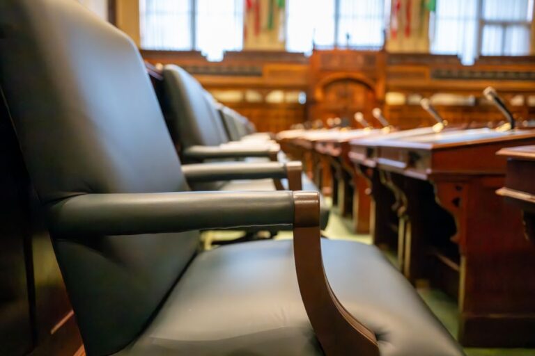 Empty leather chair in a formal government chamber; wooden desks and microphones visible in the background.
