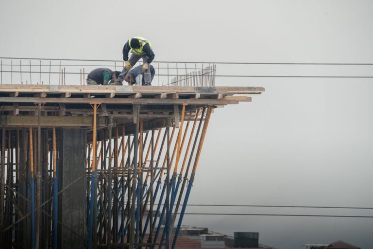 featured-35300835 - Awesim Building Consultants Construction workers in safety vests lean over the edge of a partially built elevated platform, surrounded by orange and blue scaffolding against a gray sky.