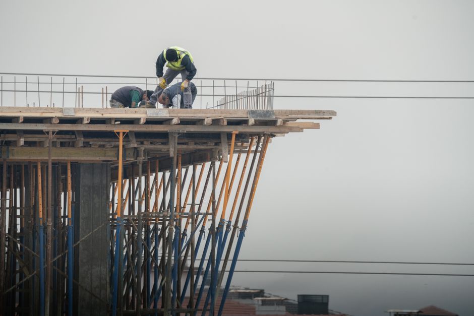 featured-35300835 - - Construction workers in safety vests lean over the edge of a partially built elevated platform, surrounded by orange and blue scaffolding against a gray sky.