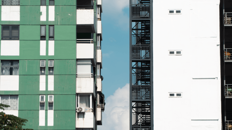 Two tall urban apartment buildings facing each other: a green striped facade on the left and a white building with a metal fire escape on the right, with blue sky between.