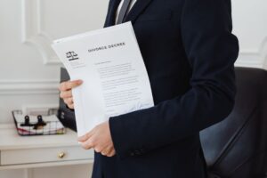 Person in a dark suit holds a divorce decree document in an office setting, the legal paper partially visible with a scales-of-justice logo.