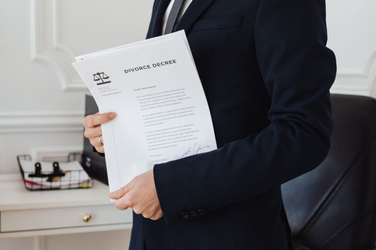Person in a dark suit holds a divorce decree document in an office setting, the legal paper partially visible with a scales-of-justice logo.