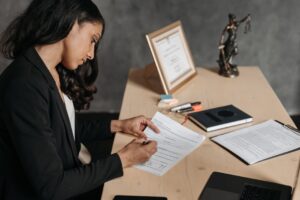 featured-8111889-1 - Professional woman in a blazer signs legal documents at a wooden desk, with a framed certificate and scales of justice nearby.