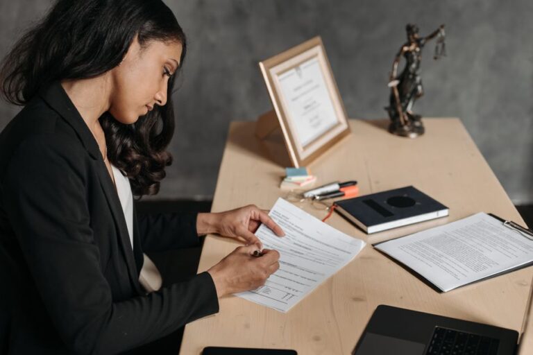 Business professional signs documents at a wooden desk with law items like a framed certificate, notebook, and a scales of justice statue in the background.