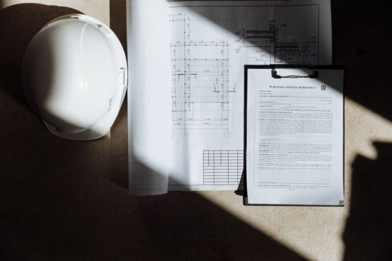 White hard hat, rolled blueprints, and a clipboard with a contract on a construction site floor, signaling project planning and safety.