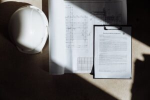 White construction helmet, rolled blueprints, and a clipboard with a purchase and sale agreement on a concrete floor.