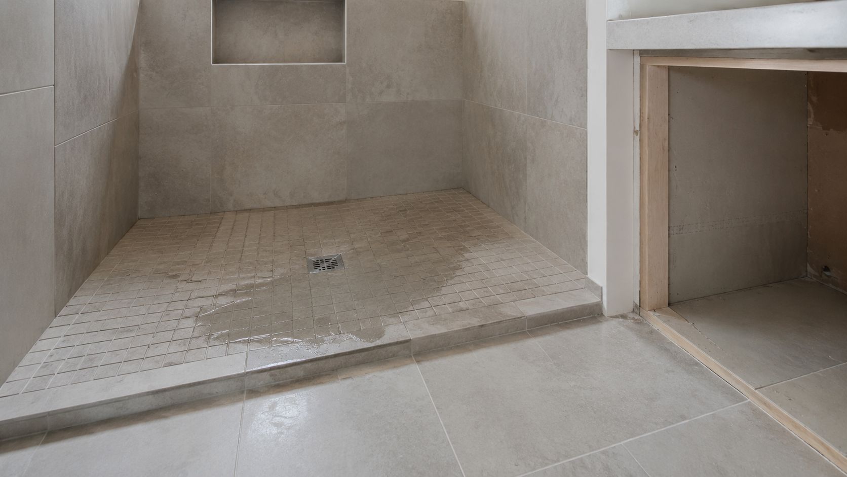 A modern shower stall with grey tiling and a small square drain under construction in a bathroom.