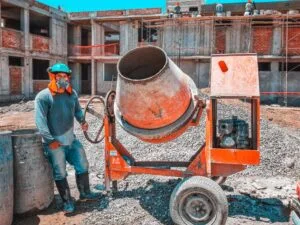 Construction worker in PPE stands beside a red portable concrete mixer on an unfinished building site with brick walls behind him.