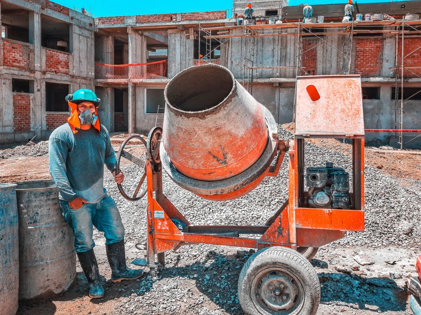 featured-6082416 - Awesim Building Consultants - Awesim Building Consultants Construction worker in PPE stands beside a red portable concrete mixer on an unfinished building site with brick walls behind him.