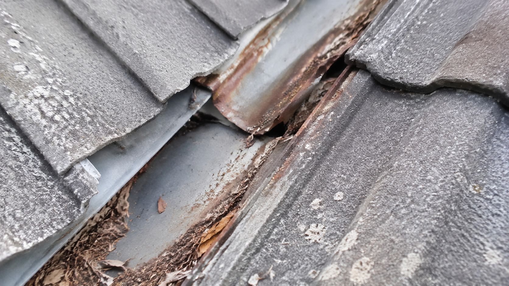 A close-up view of a deteriorating metal roof valley showing rust, debris, and damaged roof tiles.