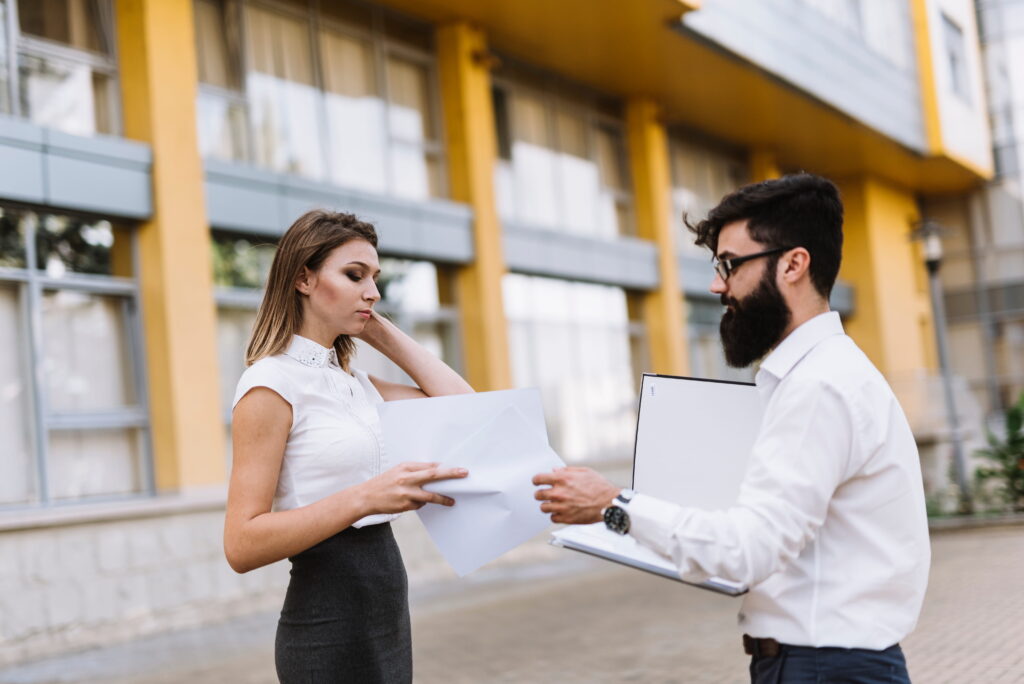 Two professionals in business attire exchange documents outdoors in front of a modern yellow-and-glass building.